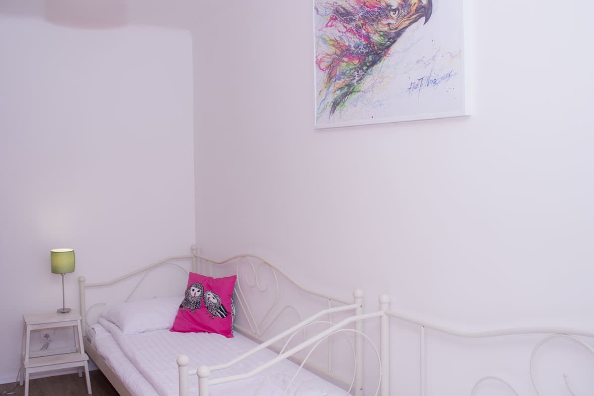 Una cama de metal blanca con un cojín rosa y una mesita de noche con una lámpara.