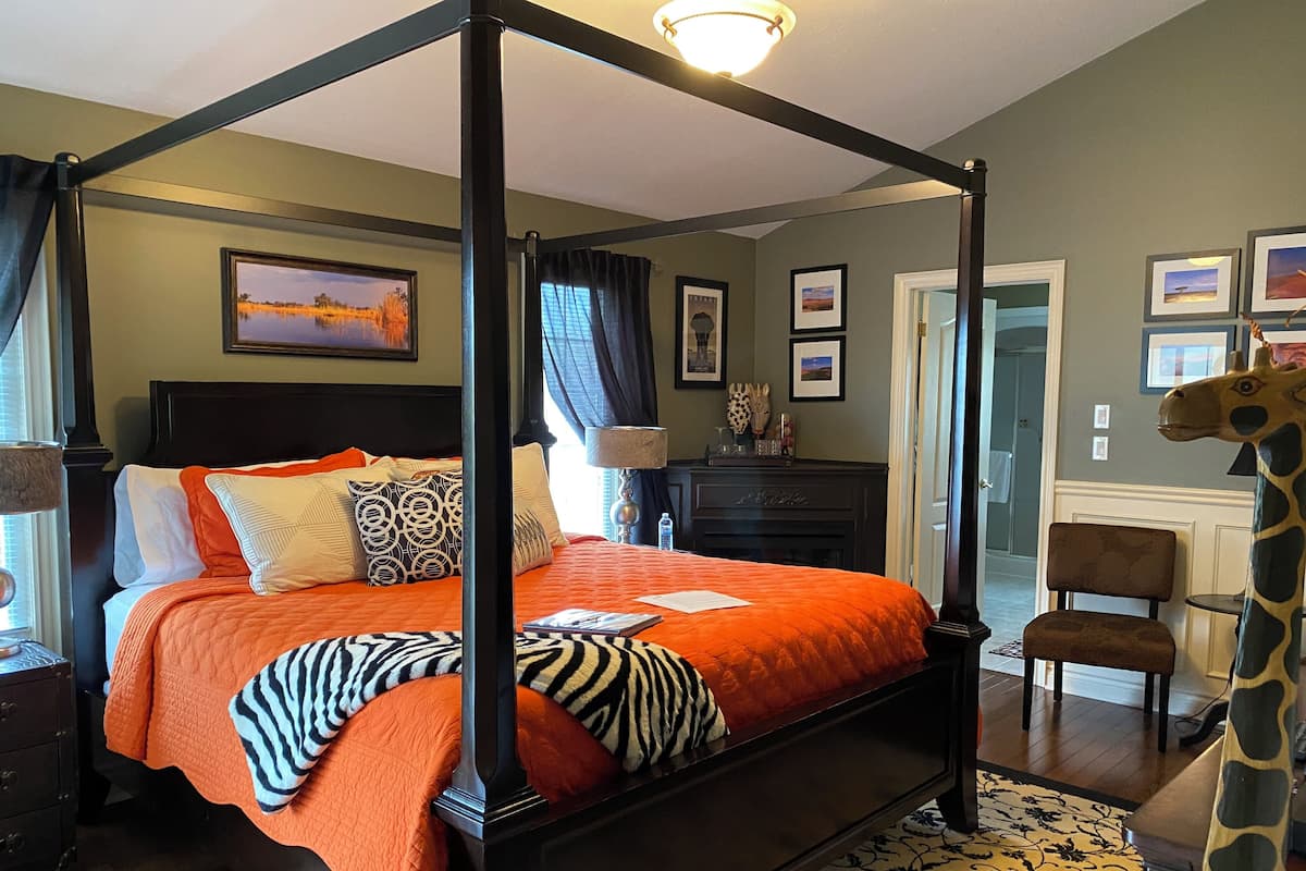 A four-poster bed with an orange bedspread, a zebra print throw, and multiple pillows. There is a dark wooden dresser to the left, a lamp, and a giraffe statue to the right.