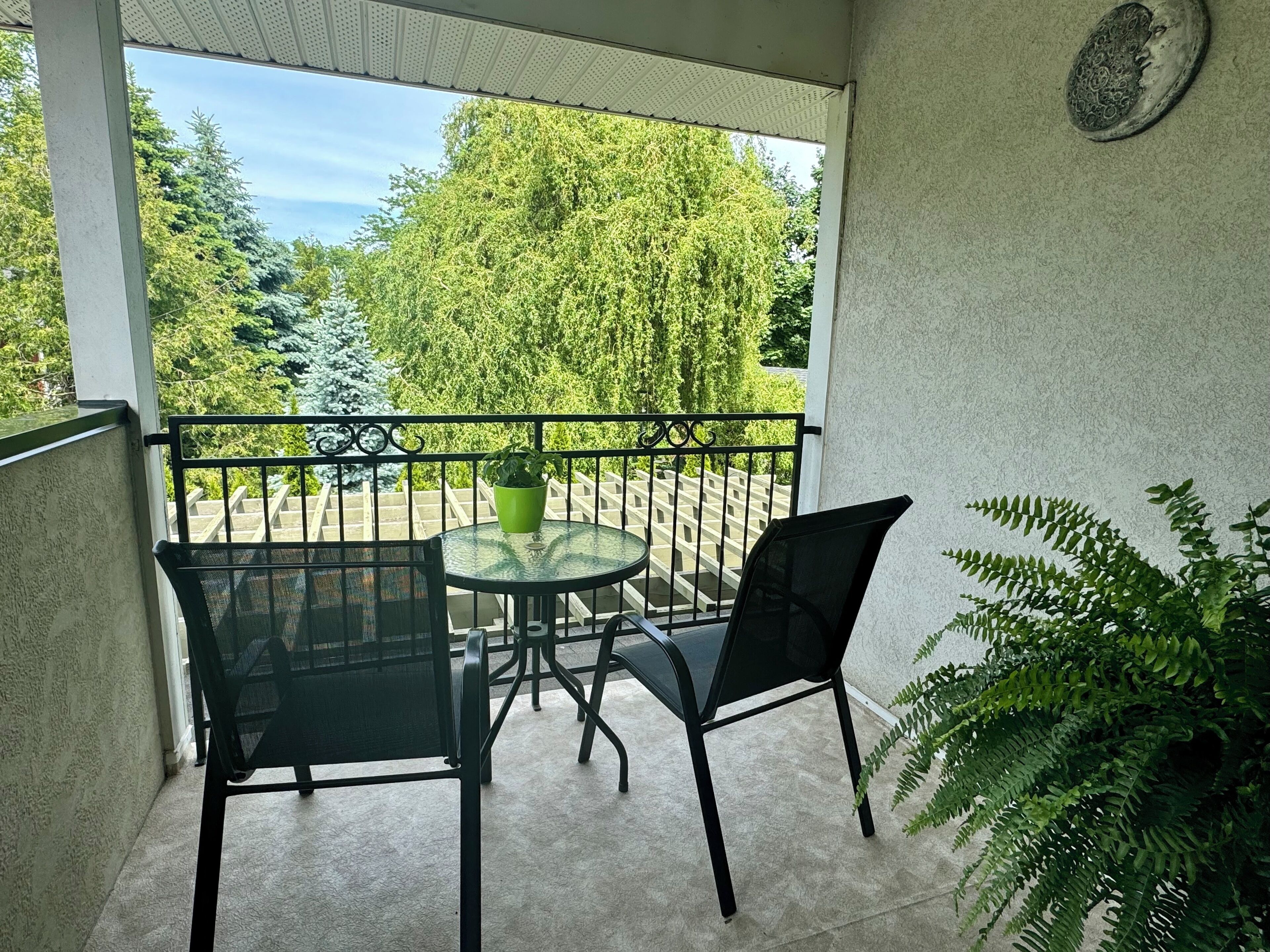 Ein Balkon mit Glastisch und Stühlen, einem Blumentopf und Blick auf einen Garten mit Bäumen und einem Holzzaun.