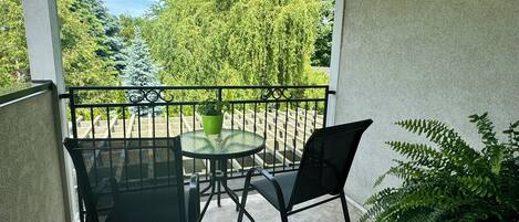 Ein Balkon mit Glastisch und Stühlen, einem Blumentopf und Blick auf einen Garten mit Bäumen und einem Holzzaun.