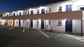 A row of single-story motel rooms with individual doorways and windows, a parking lot in front, and a clear sky at dusk. - Budget Inn Motel Suites (Somers Point)