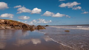 Una playa con aguas tranquilas, costa rocosa y un cielo azul despejado con nubes.