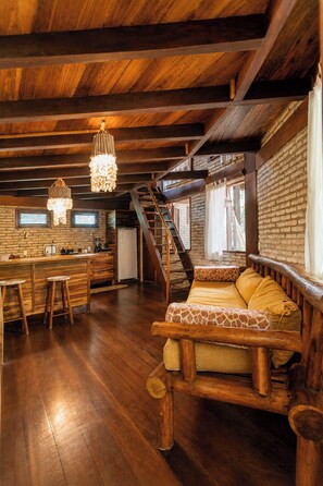 Un interior rústico con una escalera de madera, un sofá con cojines de estampado de jirafa, y una zona de cocina con pared de ladrillo y vigas de madera.