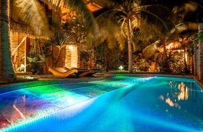 Una piscina de borde curvo, rodeada de palmeras y una estructura con techo de paja.