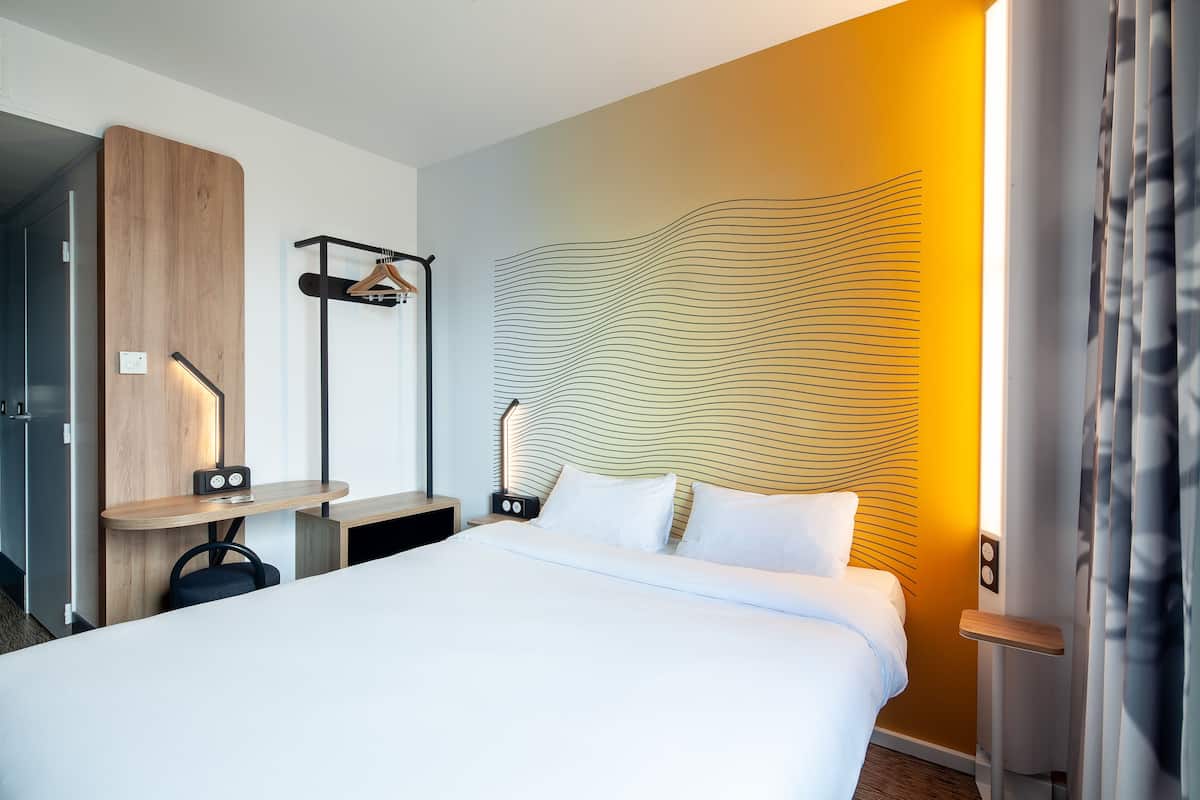 Een moderne hotelkamer met een groot bed, een houten garderobe, een bureau met stoel, en een opvallend wandontwerp.
