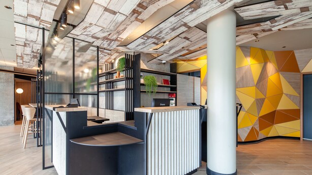 Een moderne receptiezone met een houten receptiebalie, een zithoek met stoelen en een wand met een geometrisch patroon.