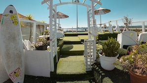 A white archway with umbrellas, potted plants, and surfboards in a sunny outdoor setting. - Sunset Destination Hostel (Lisbon)