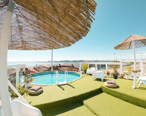 A rooftop pool area with a clear view of the sea, surrounded by white lounge chairs and a thatched umbrella.