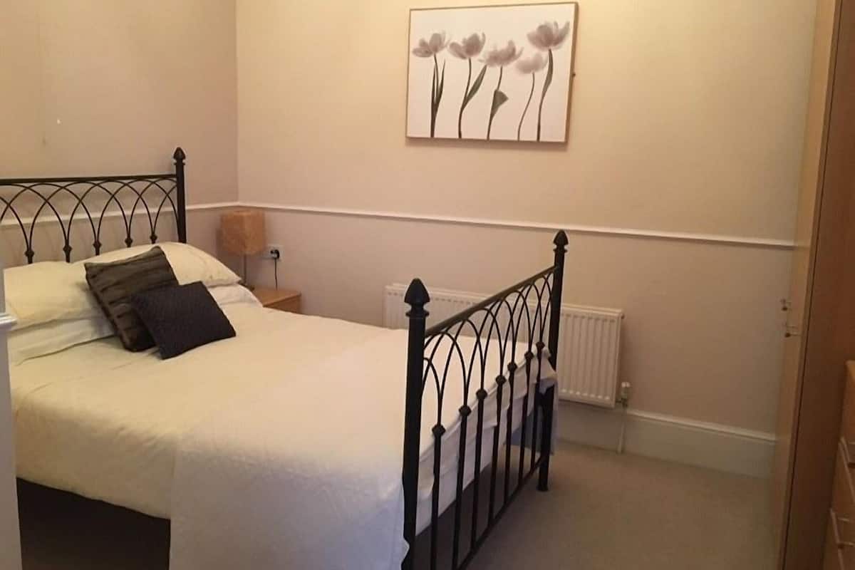 A bedroom with a bed, a nightstand, a lamp, a radiator, and a wall-mounted picture of flowers.
