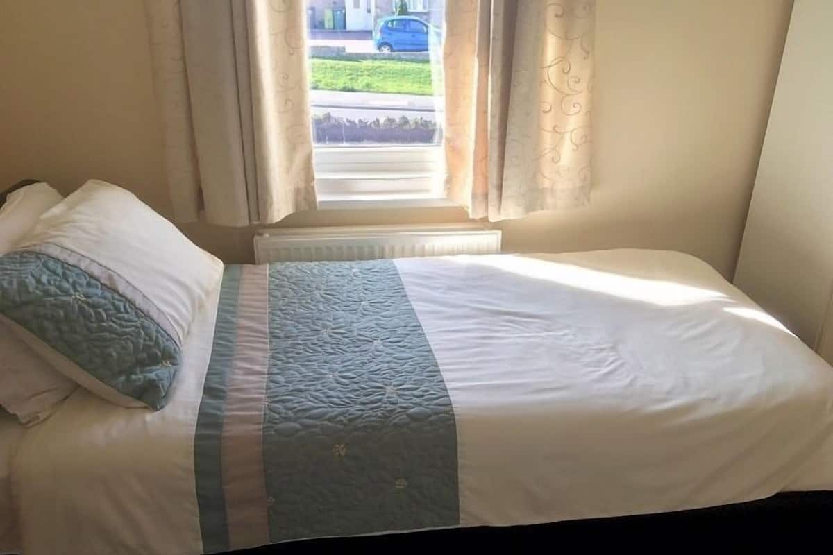 A single bed with a white and blue quilt, a pillow, and a window with curtains.
