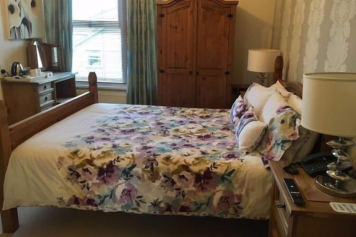 A hotel room with a wooden bed, floral bedspread, a wooden wardrobe, a desk with a lamp, and a view of the outdoors through the window.