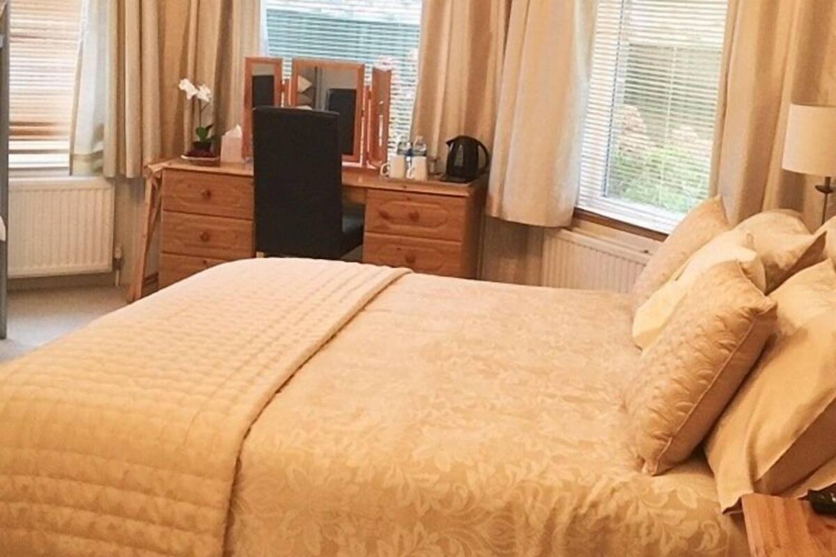 A neatly arranged bedroom with a large bed, a wooden dresser, a desk with a chair, and a window with curtains.