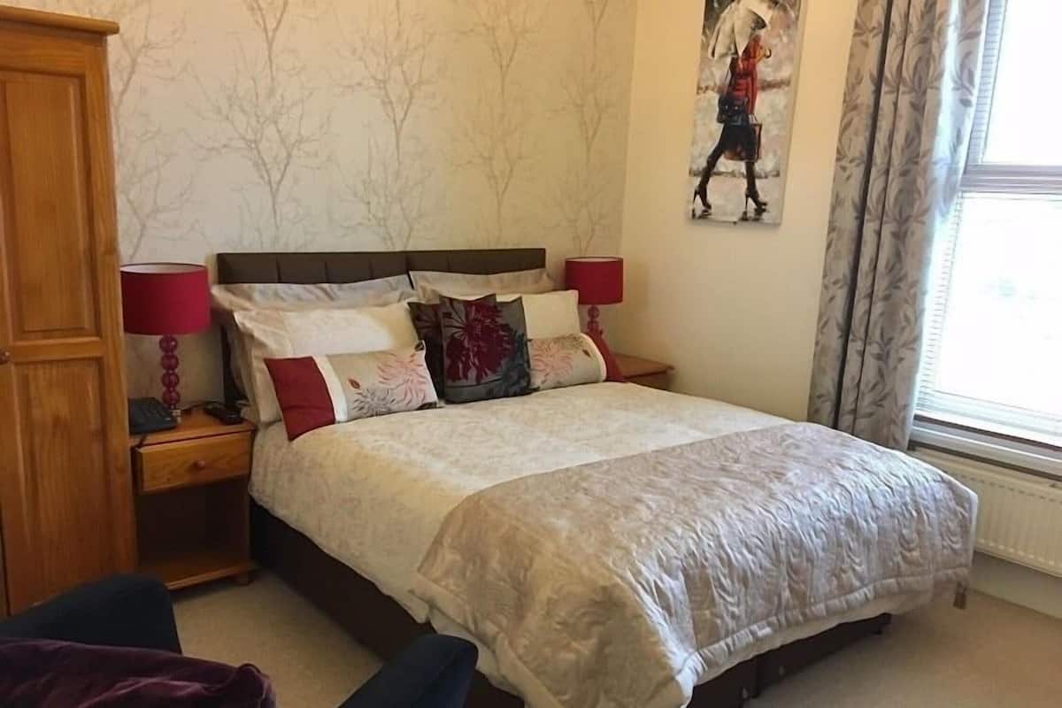 A neatly made bed with a patterned bedspread, two bedside tables with lamps, a wooden wardrobe, and a window with curtains.