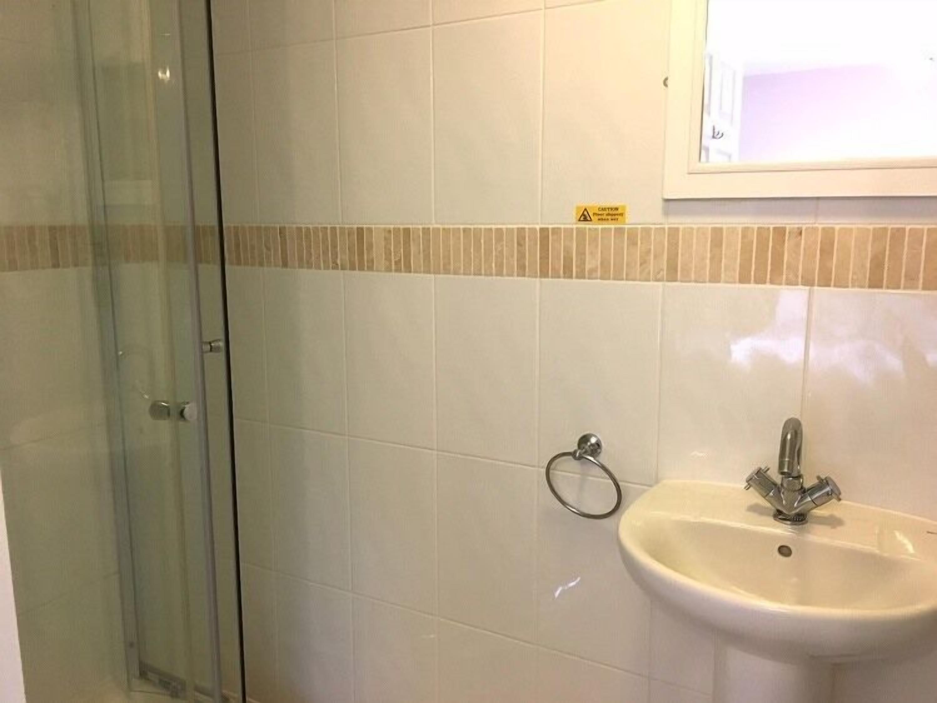 Un baño con cabina de ducha de vidrio, un lavamanos con grifo y una pared con azulejos y un letrero de precaución.