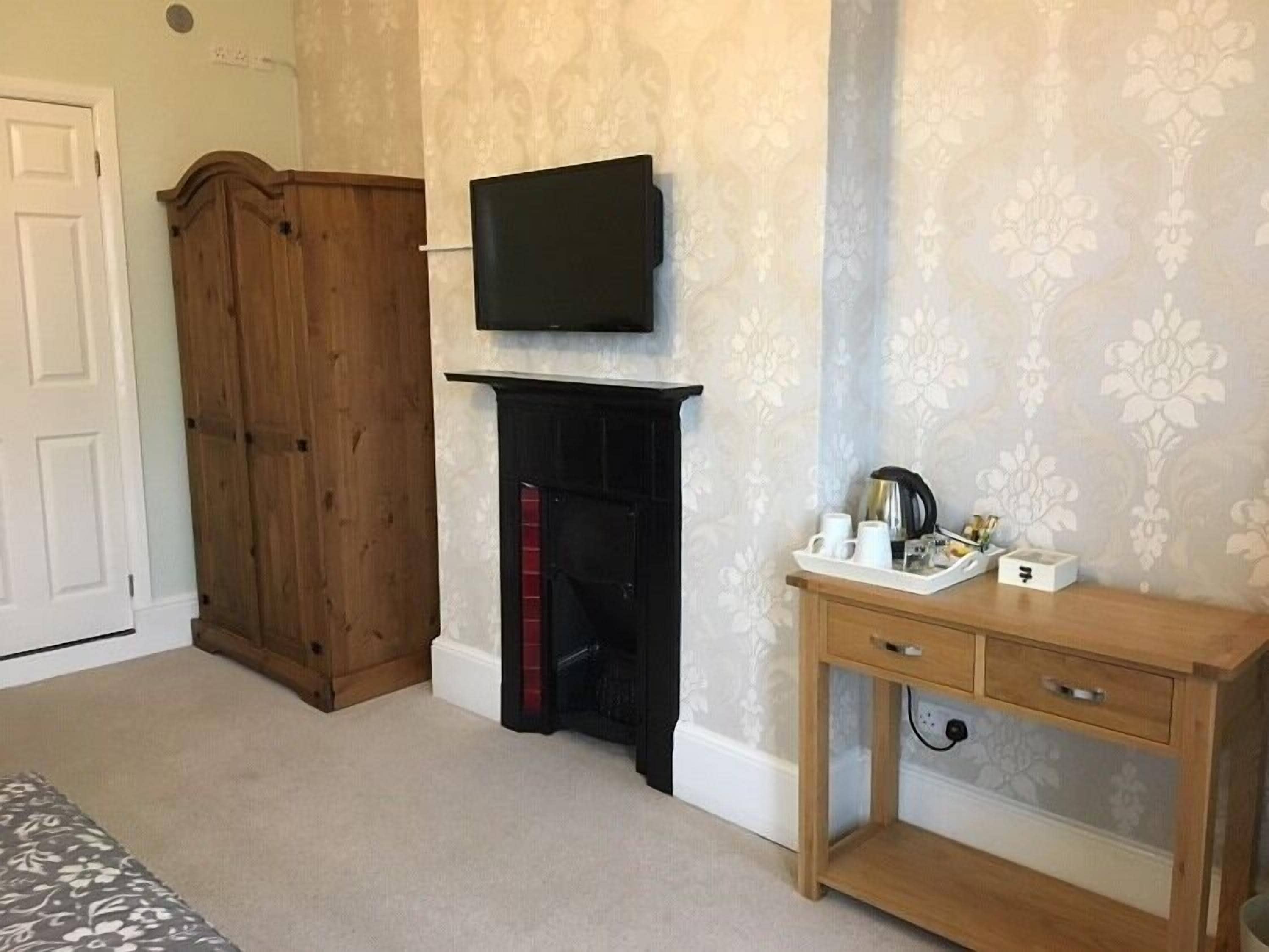 A room with a wooden wardrobe, a fireplace with a black mantel, a television mounted above it, and a wooden console table with a tray of items.