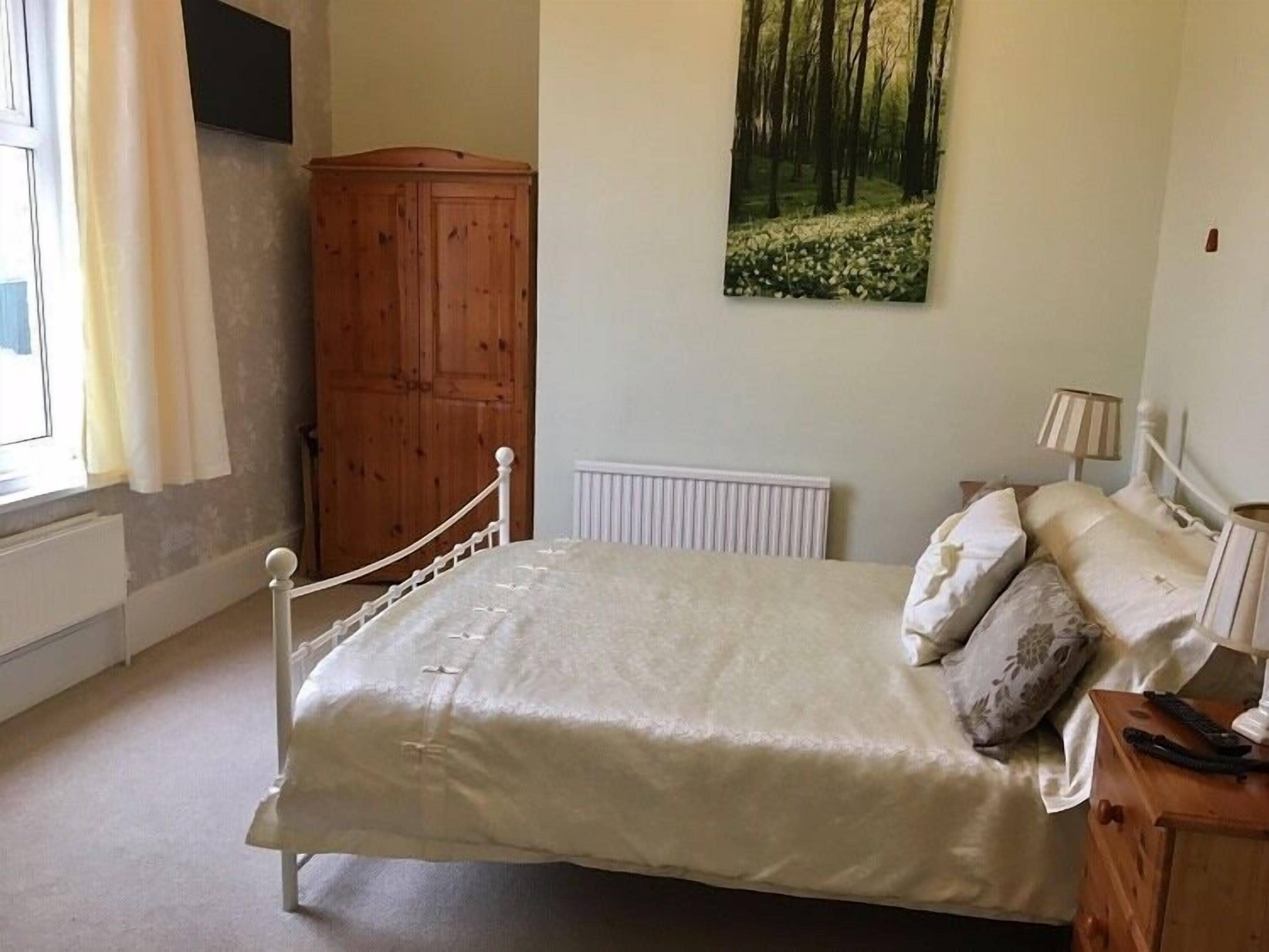 Une chambre avec un lit simple, une armoire en bois, un radiateur, une fenêtre avec des rideaux et un tableau sur le thème de la forêt accroché au mur.