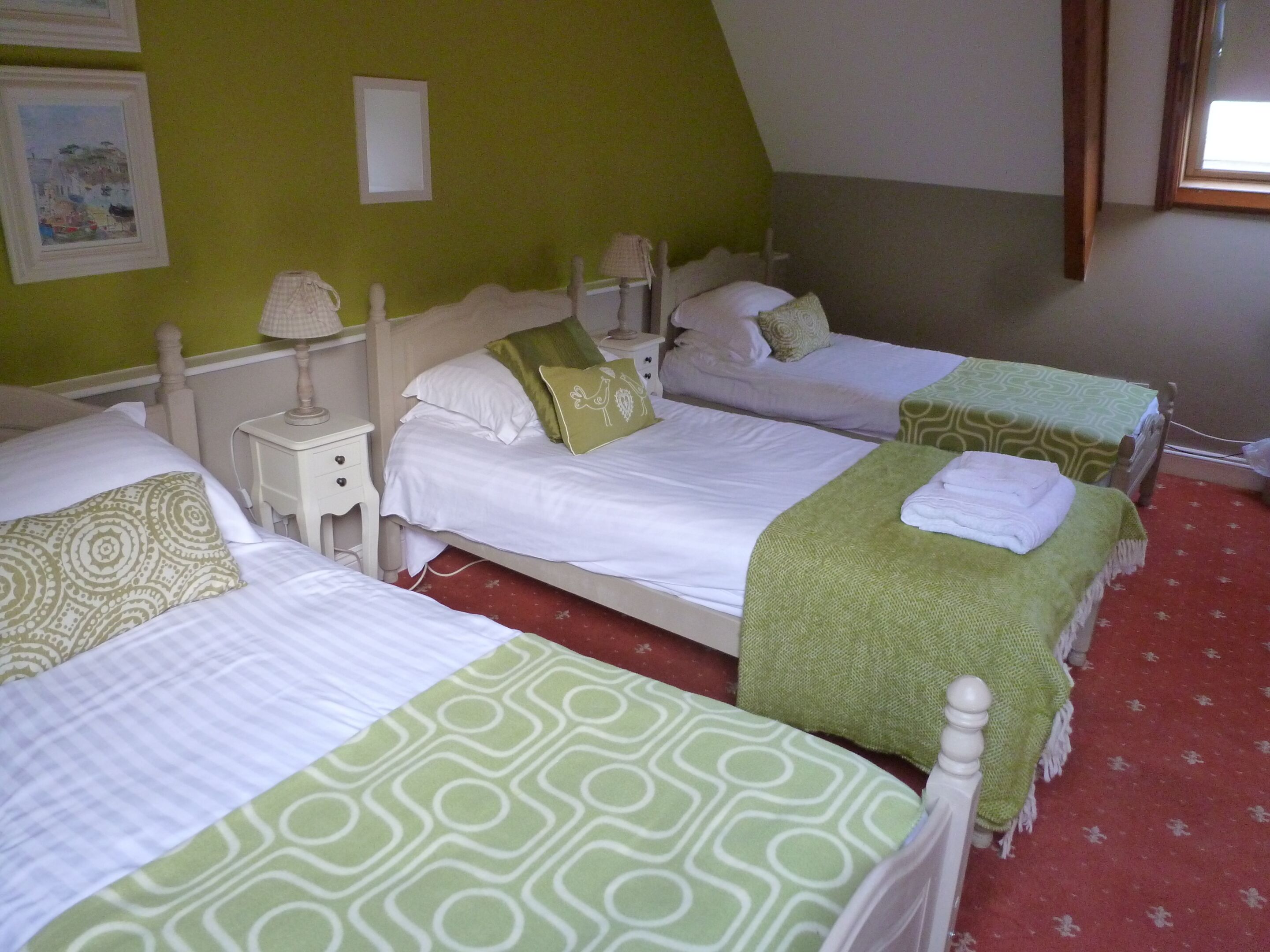 A hotel room with three beds, each with a green patterned bedspread. There are two bedside tables with lamps, and the walls are painted green.