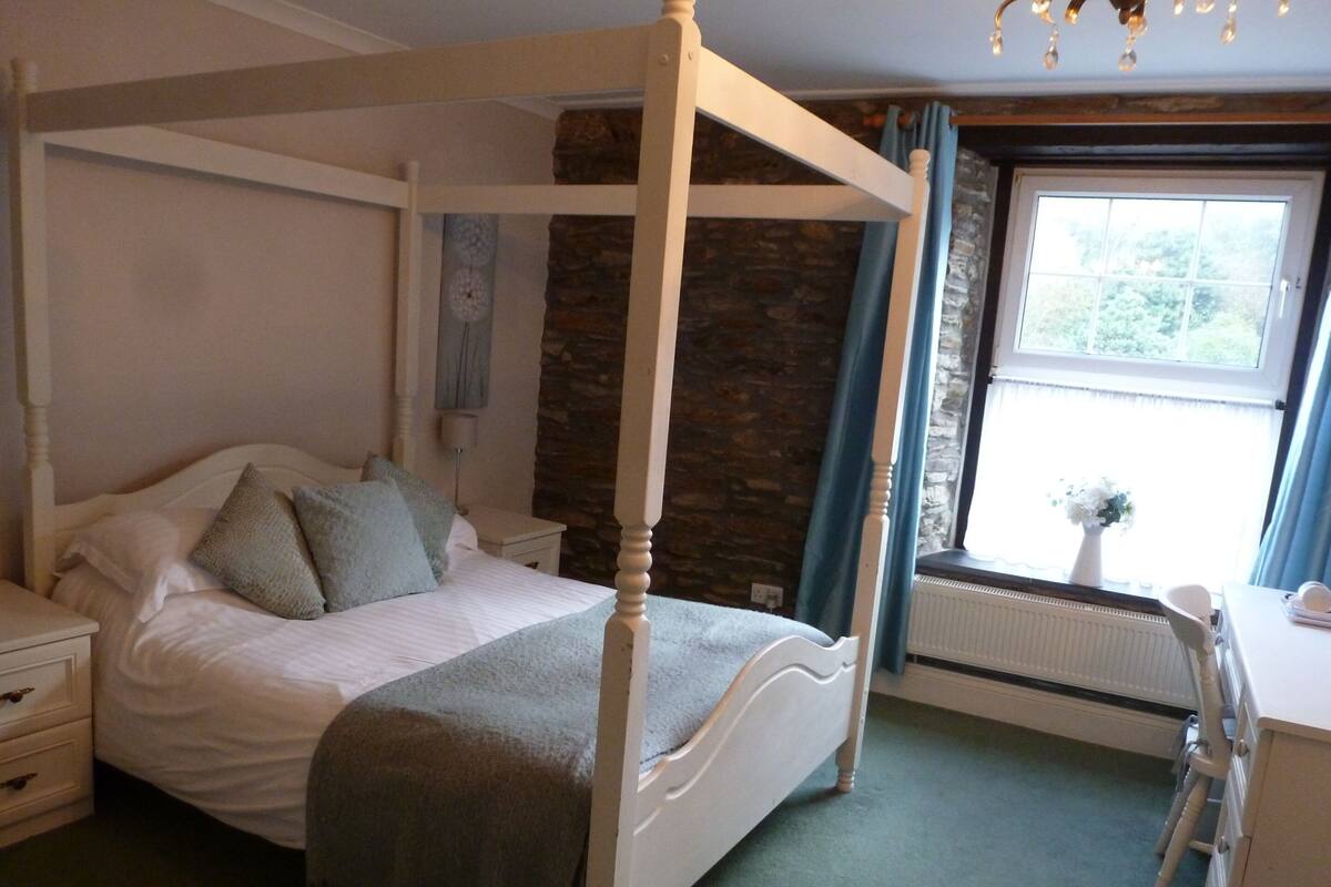 A four-poster bed with a stone wall backdrop, a bedside table, and a window with curtains.