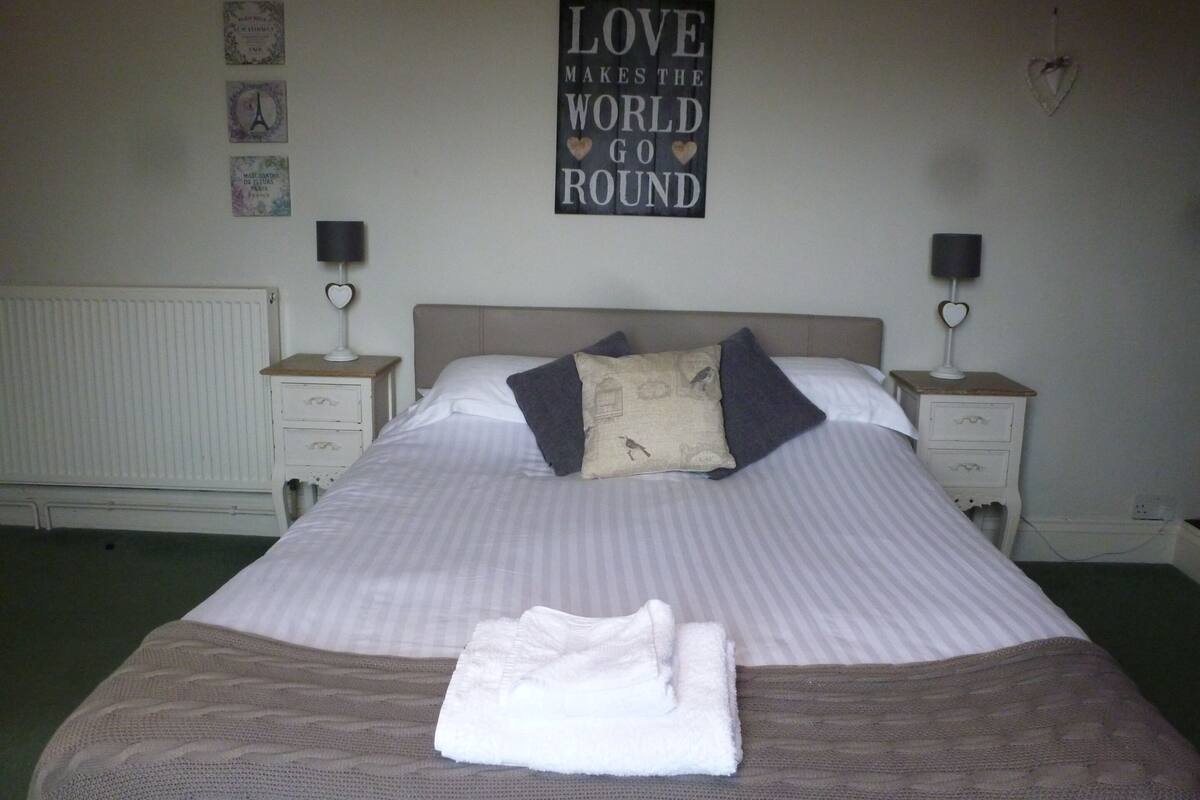 A neatly made bed with a folded towel, two bedside tables with lamps, a radiator, and a wall with a motivational quote.