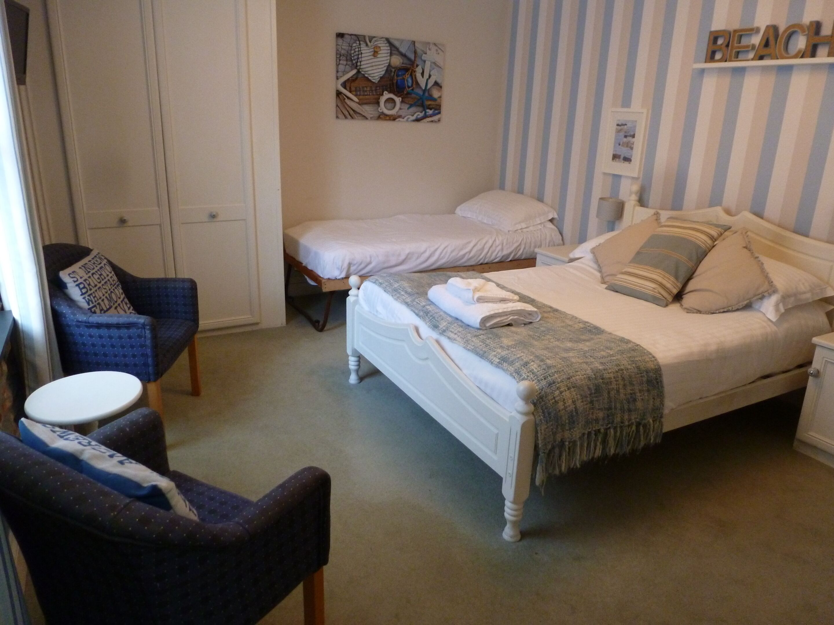 Ein Schlafzimmer mit zwei Einzelbetten, einem Sessel, einem kleinen runden Tisch und einem an der Wand befestigten Regal mit der Aufschrift 'BEACH'.
