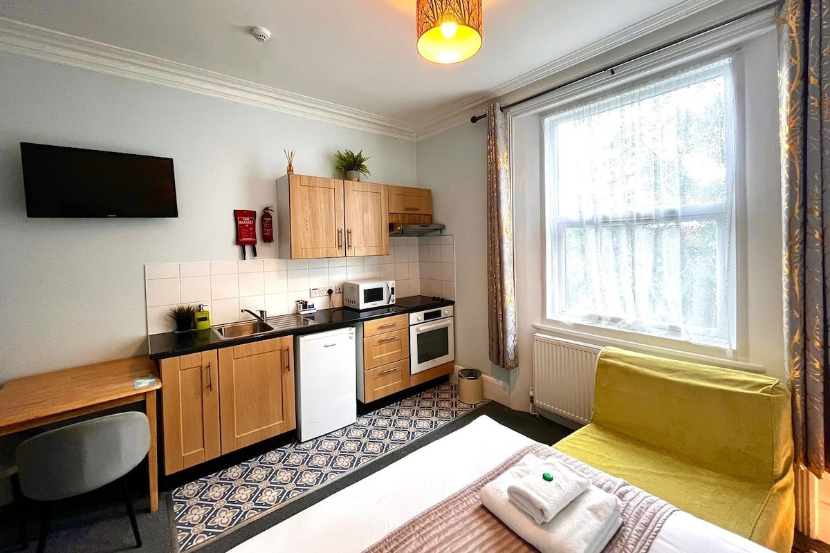 Een compacte hotelkamer met een kitchenette, een bed met een gele hoofdeinde, een aan de muur bevestigde televisie en een raam met gordijnen.