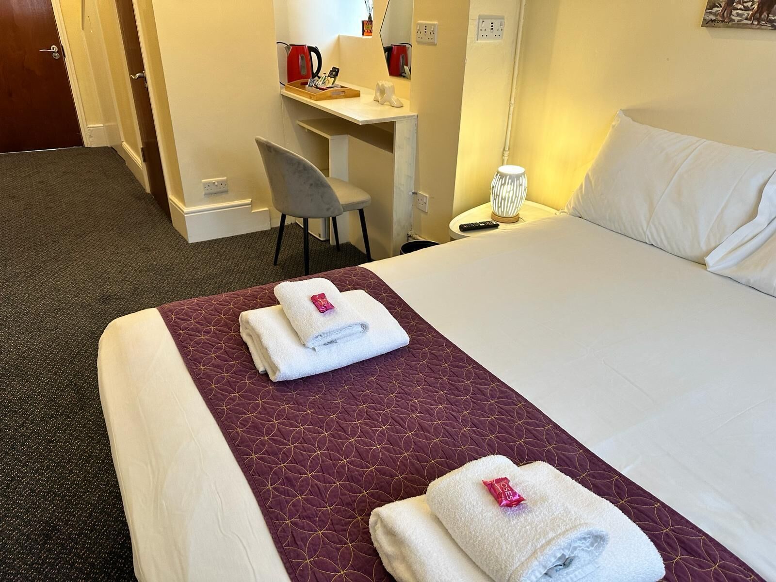A hotel room with a bed, a desk, a chair, and a nightstand with a lamp.