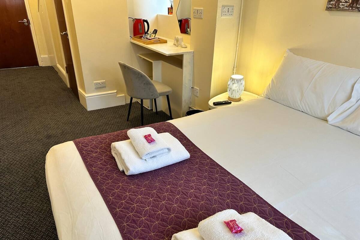 A hotel room with a bed, a desk, a chair, and a nightstand with a lamp.