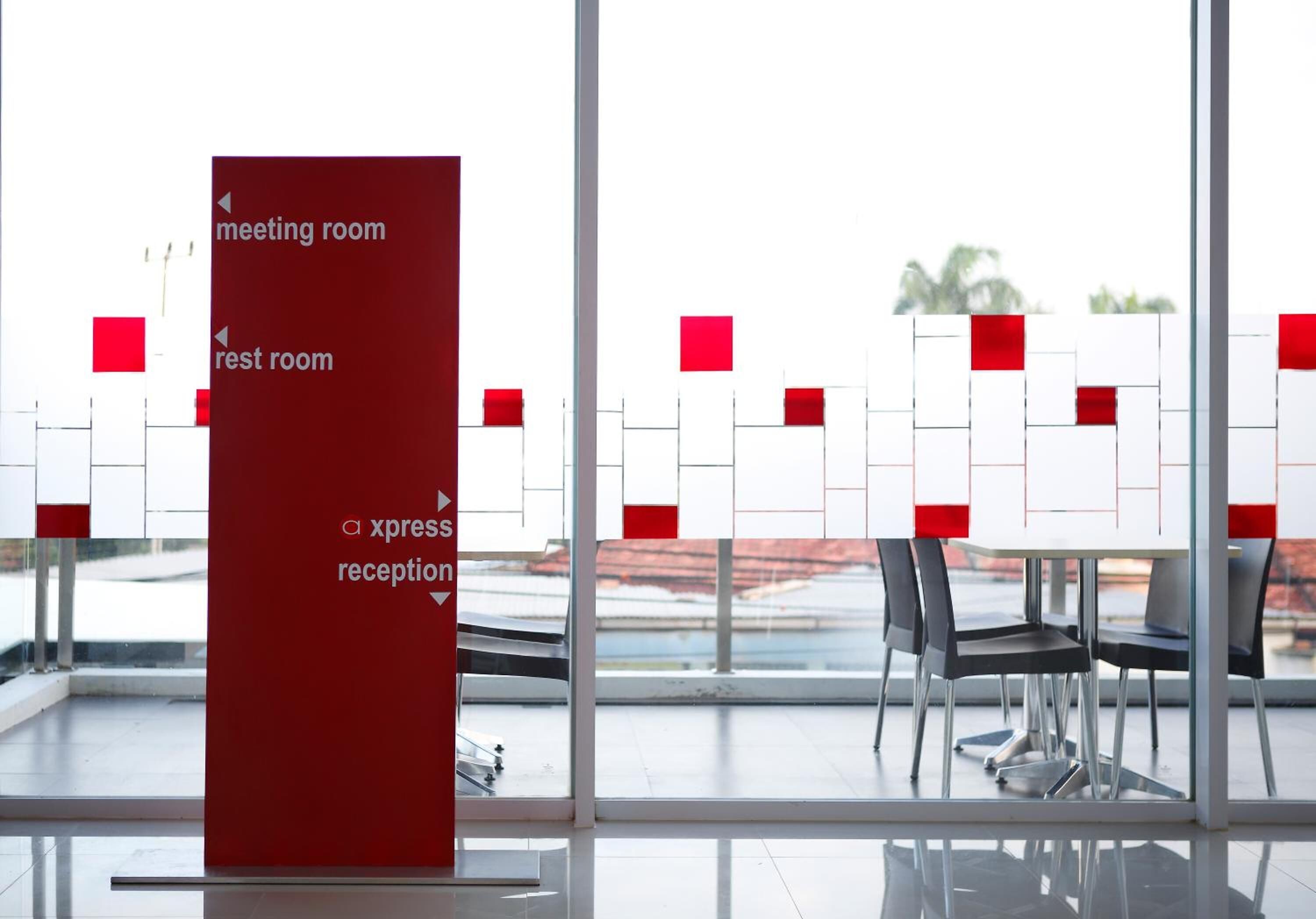 A modern hotel lobby with a glass partition, a red sign indicating meeting, rest, and reception areas, and a view of a red building outside.
