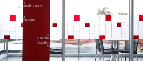 A modern hotel lobby with a glass partition, a red sign indicating meeting, rest, and reception areas, and a view of a red building outside.