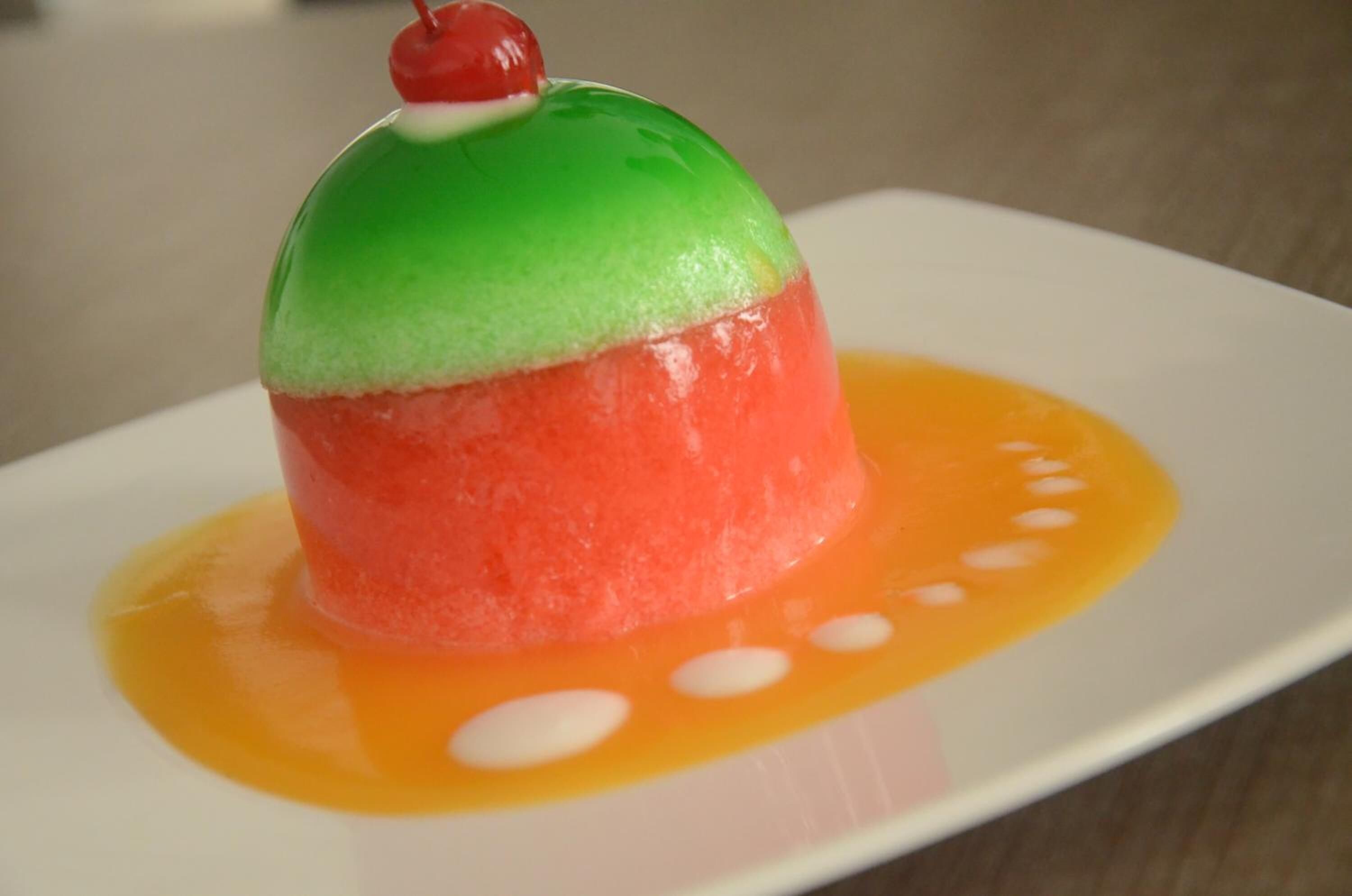 A dessert on a white plate with a green and red dome, a cherry on top, and orange sauce with white dots.