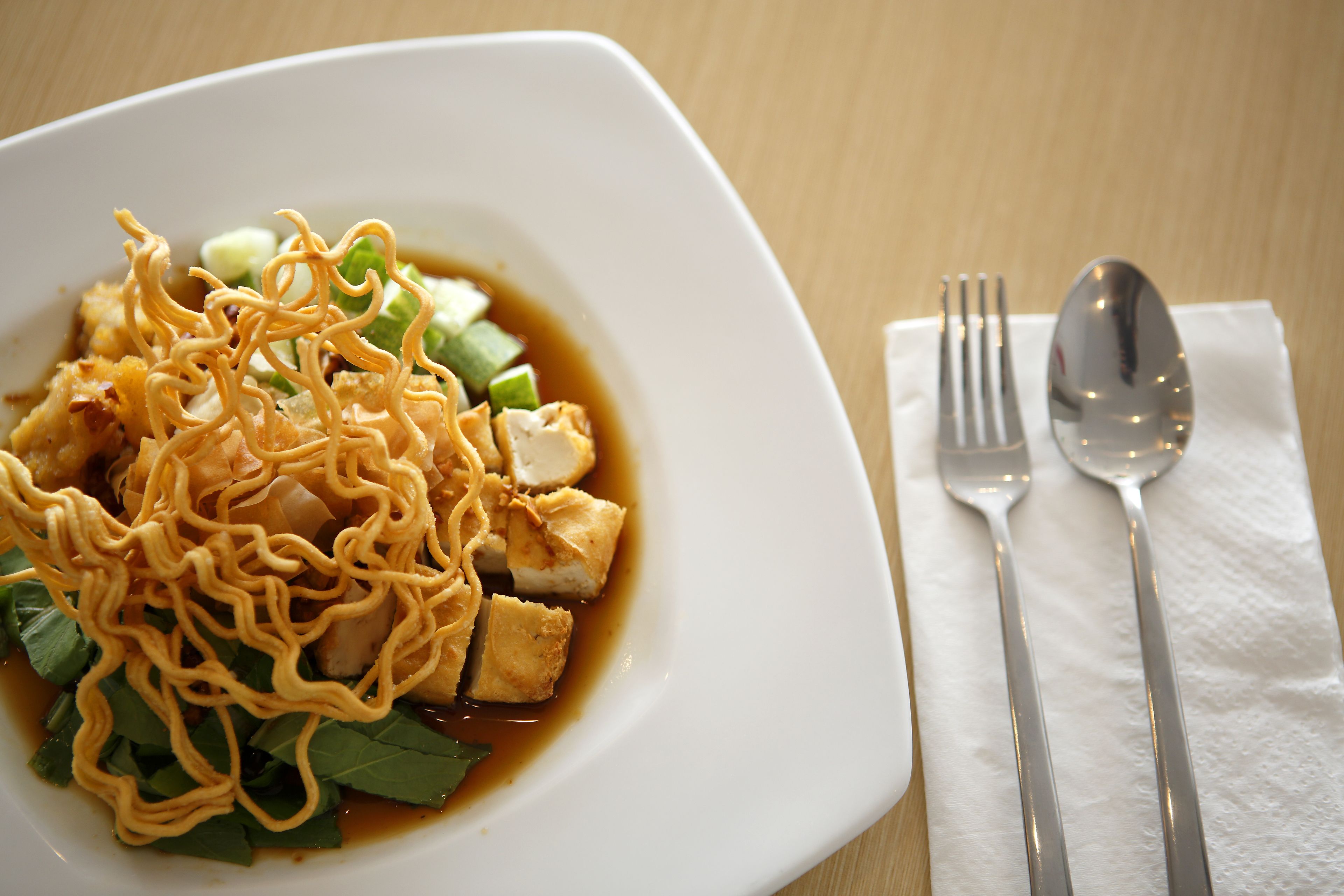 A white plate with noodles, tofu, and vegetables on a wooden table.