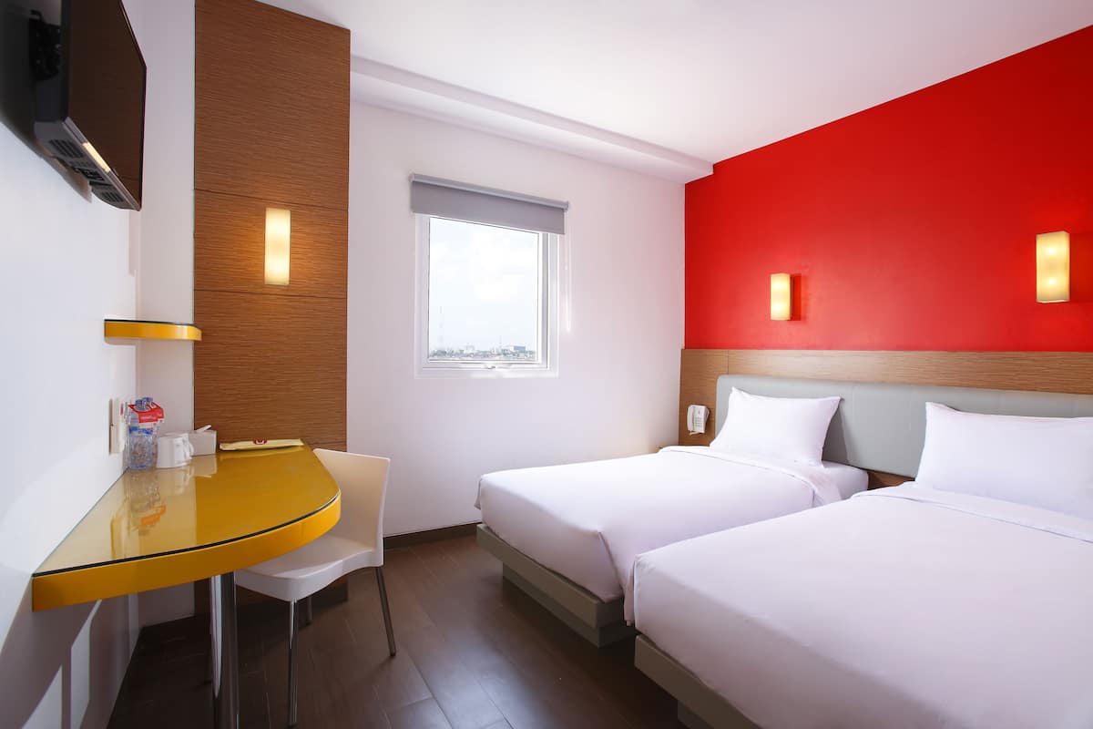 A hotel room with two beds, a small table, a chair, a window, and a red accent wall.