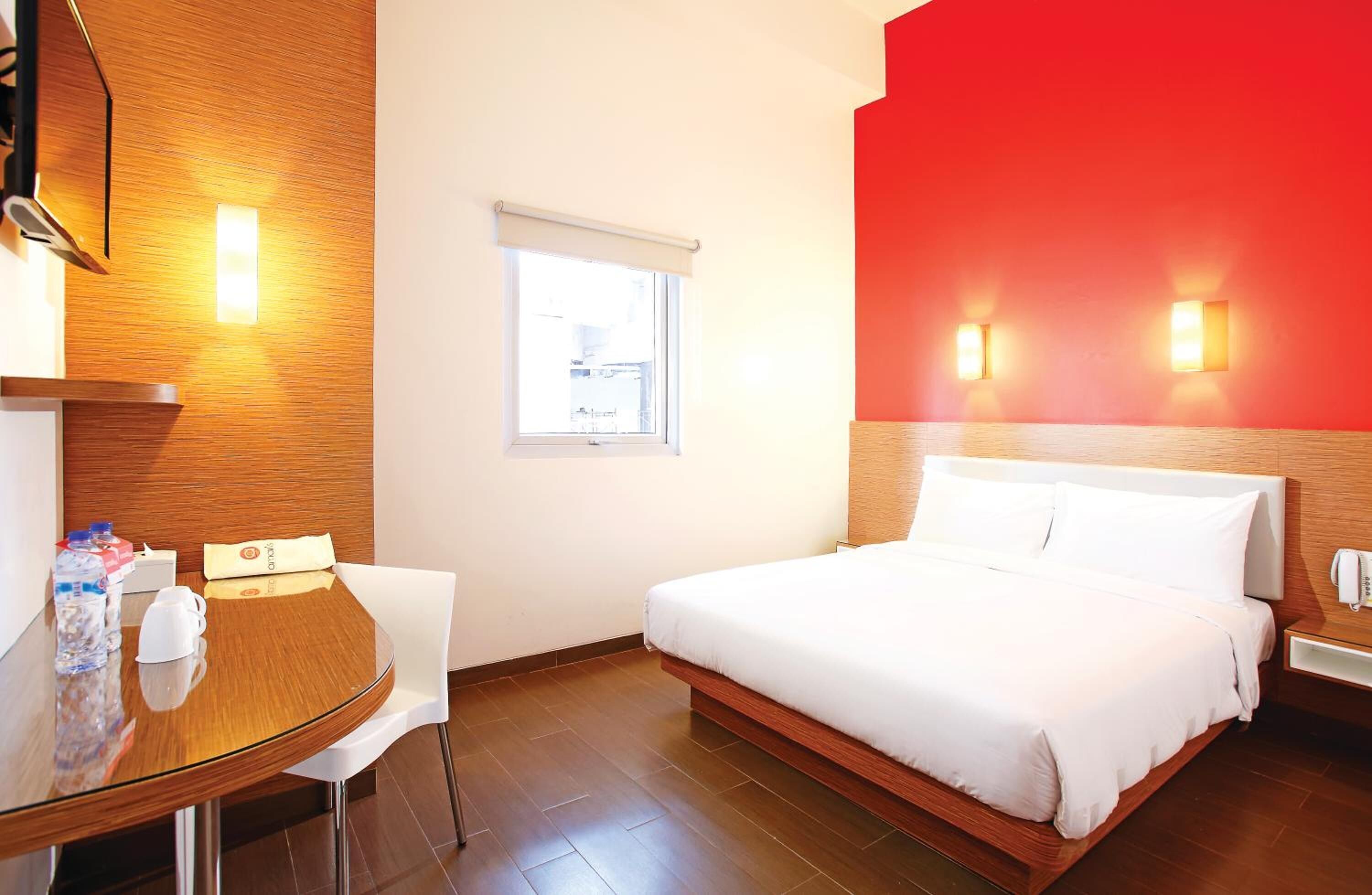 Quarto de hotel com cama, uma pequena mesa com cadeiras, janela e luminárias fixadas na parede.