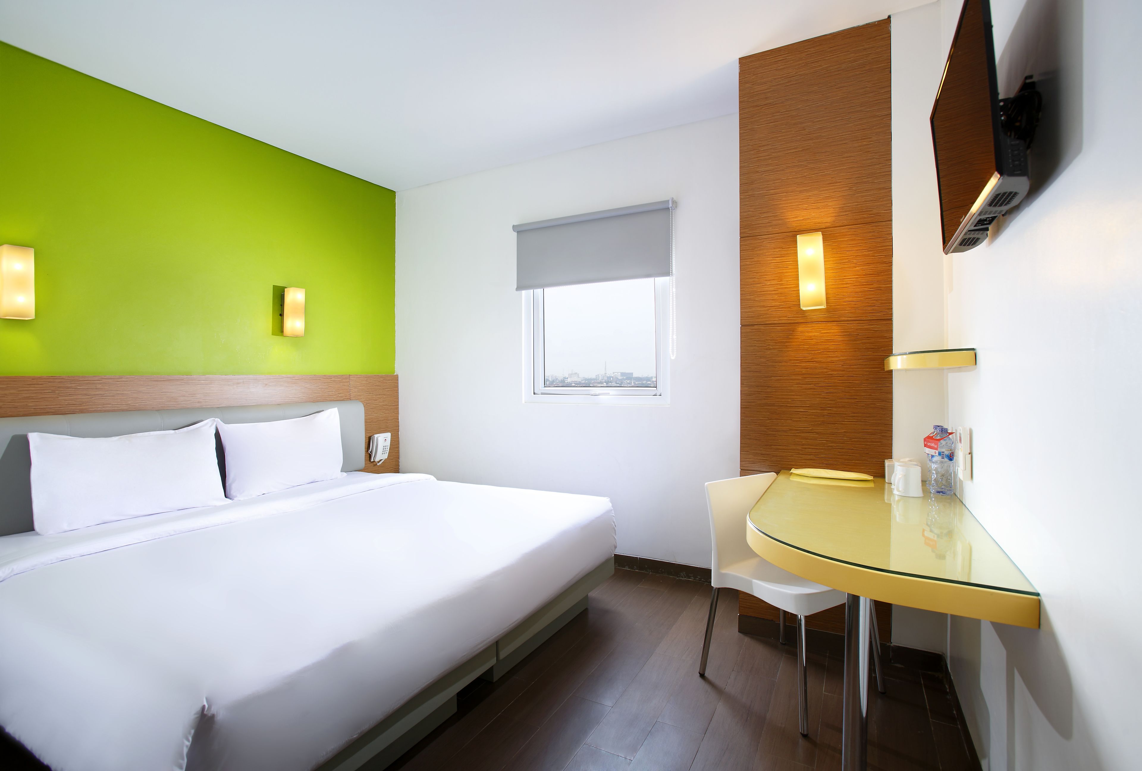 A hotel room with a bed, a glass table, a chair, a TV mounted on the wall, and a window with a blind.