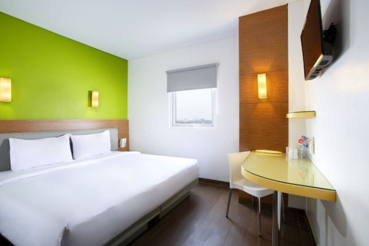 Une chambre d’hôtel comprenant un lit, un bureau avec une chaise, une fenêtre avec un store et un téléviseur fixé au mur.