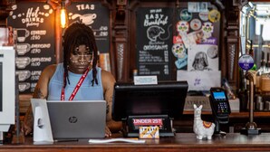 A barista at a coffee shop counter with a laptop, a payment terminal, and a small decorative llama. - PubLove The White Ferry, Victoria - Hostel (London)