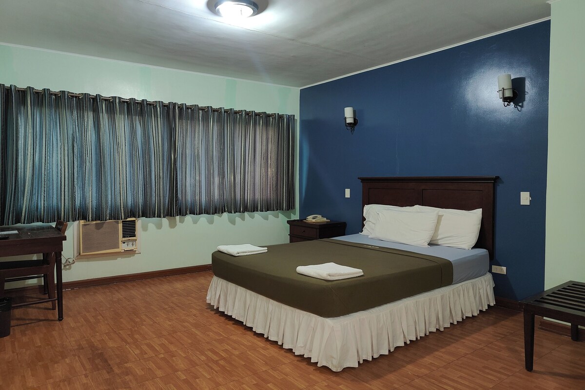 A hotel room with a bed, a desk, a chair, a nightstand, and a wall-mounted phone.