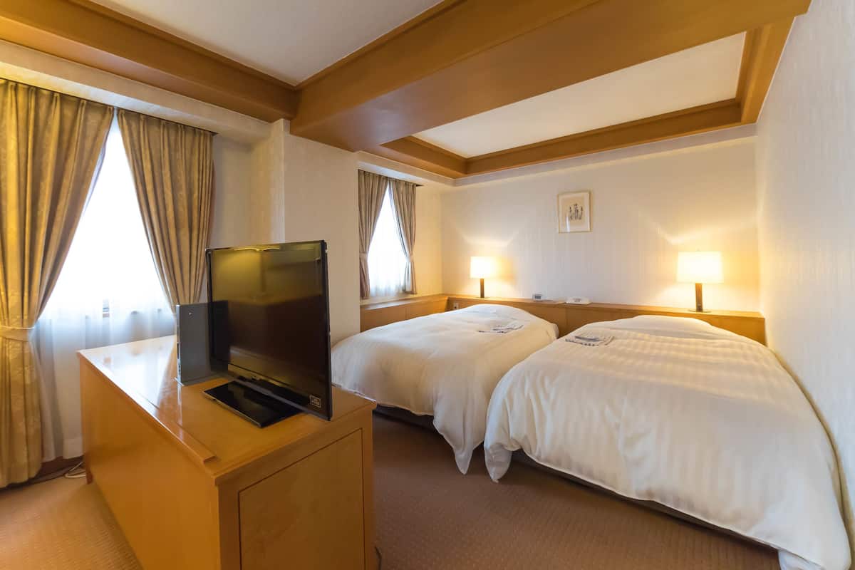 Ein Hotelzimmer mit zwei Betten, einem Schreibtisch mit Fernseher und einem Fenster mit Vorhängen.