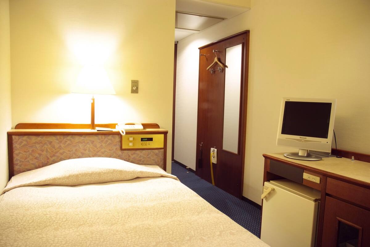 Ein Hotelzimmer mit Bett, Schreibtisch, Computerbildschirm, Lampe und Tür.