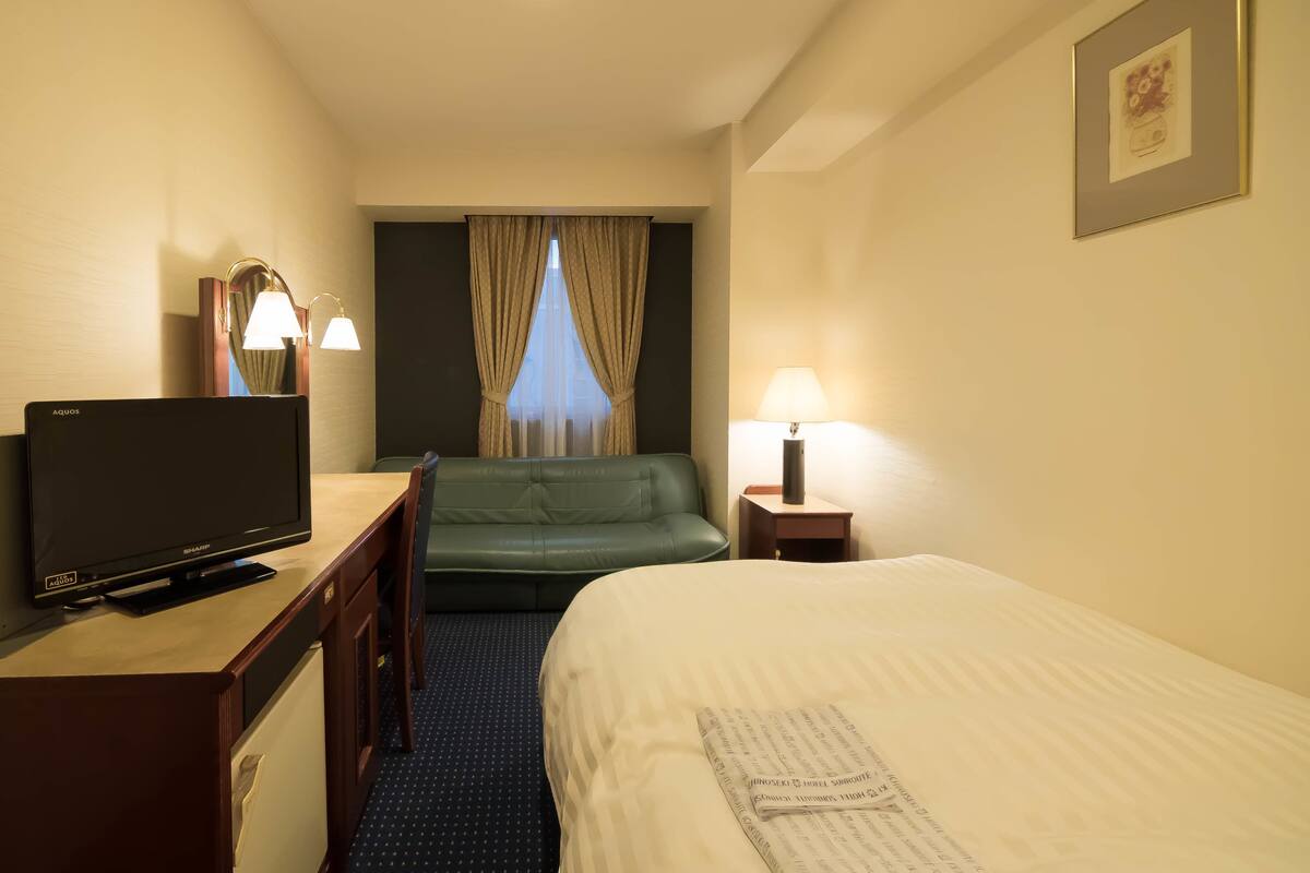 Ein Hotelzimmer mit einem Bett, einem Schreibtisch mit Fernseher, einem Sessel, einem Fenster mit Vorhängen und einer Nachttischlampe.