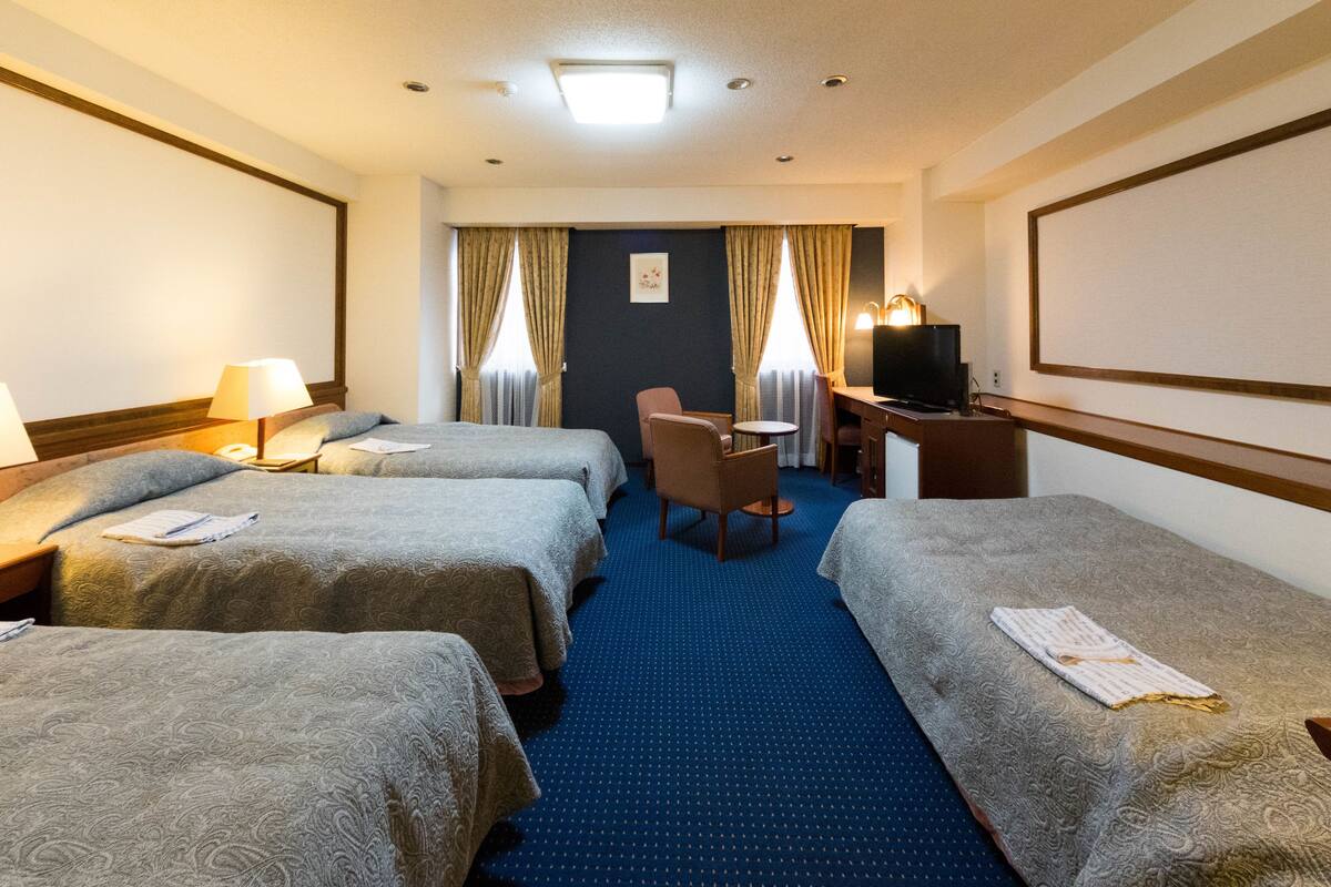 Ein Hotelzimmer mit zwei Betten, einem Schreibtisch, einem Sessel, einem Fernseher und einem großen Fenster mit Vorhängen.