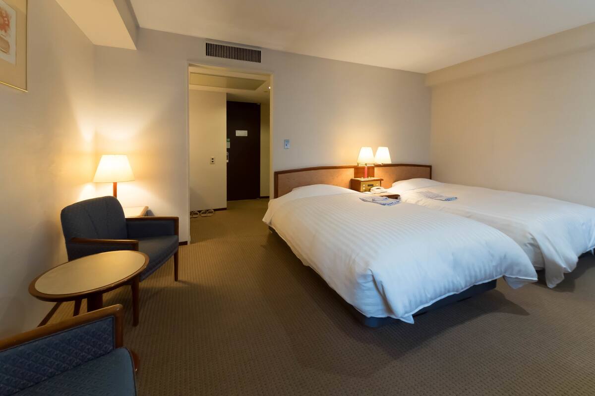 Ein Hotelzimmer mit zwei Betten, einem Schreibtisch mit Stuhl, einer Lampe und einer Tür zu einem weiteren Raum.