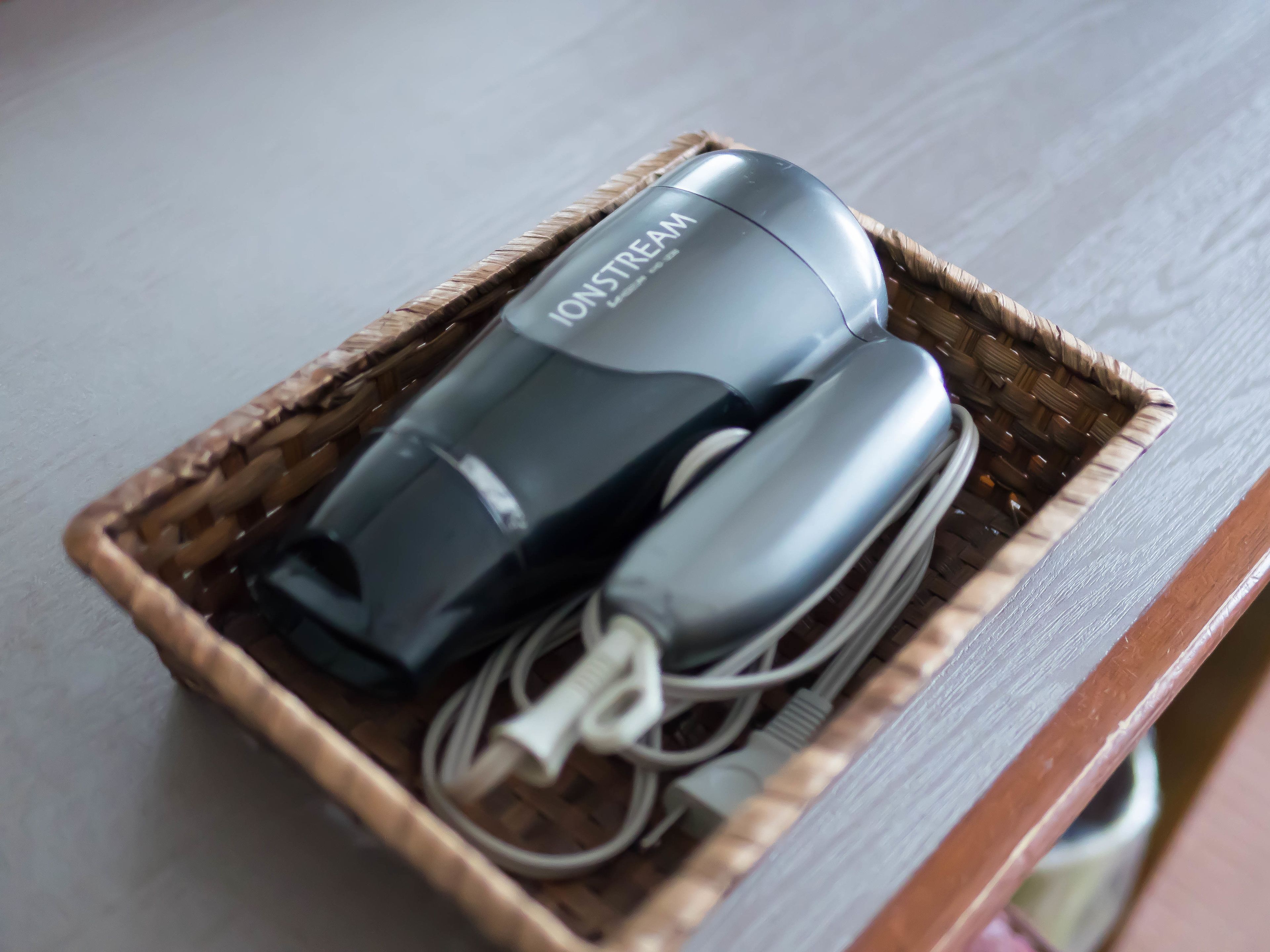 A black hair dryer and its charging cable in a woven basket on a wooden surface.