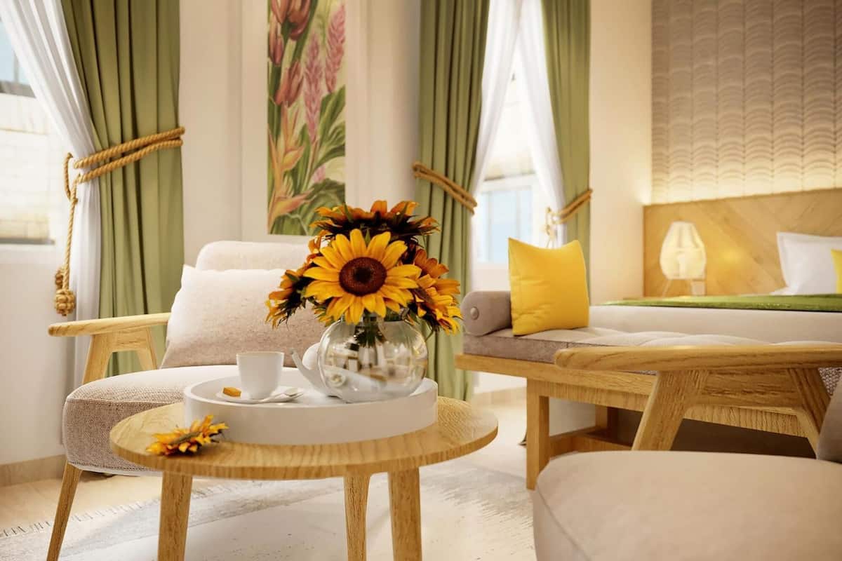 A cozy room with a round wooden table, a white sofa, and a bed with a yellow pillow. A vase with sunflowers is on the table.