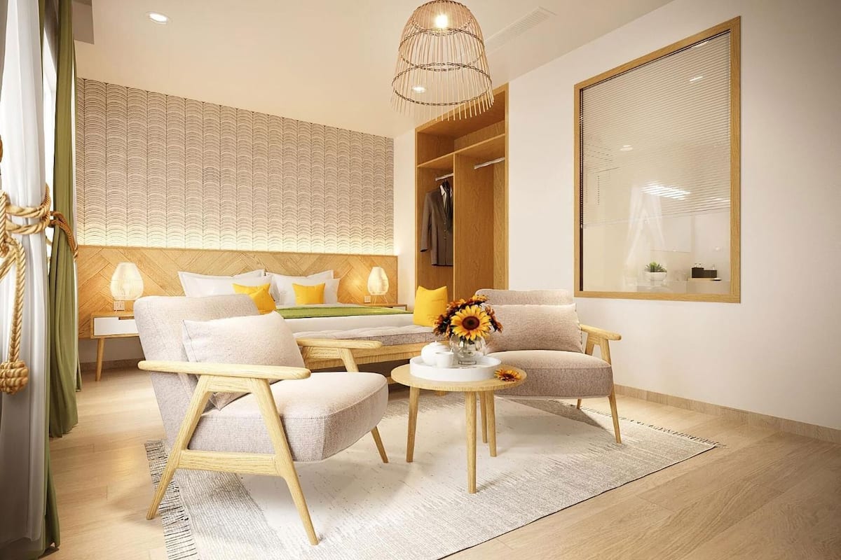 A modern hotel room with a bed, two armchairs, a small table, a hanging light fixture, a wardrobe, and a large mirror.