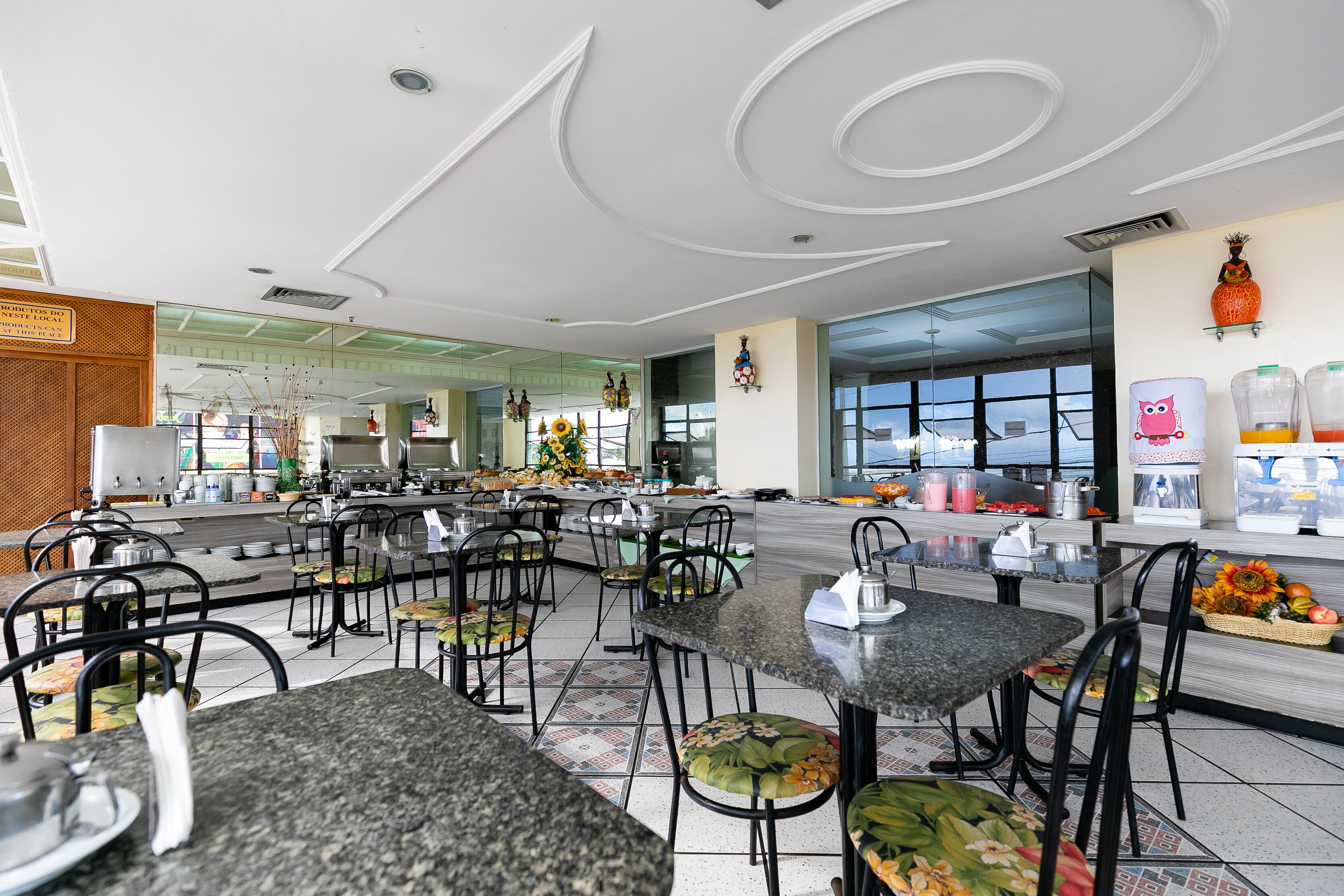 A spacious cafe with multiple tables and chairs, a large window offering a view, and a counter with various items.