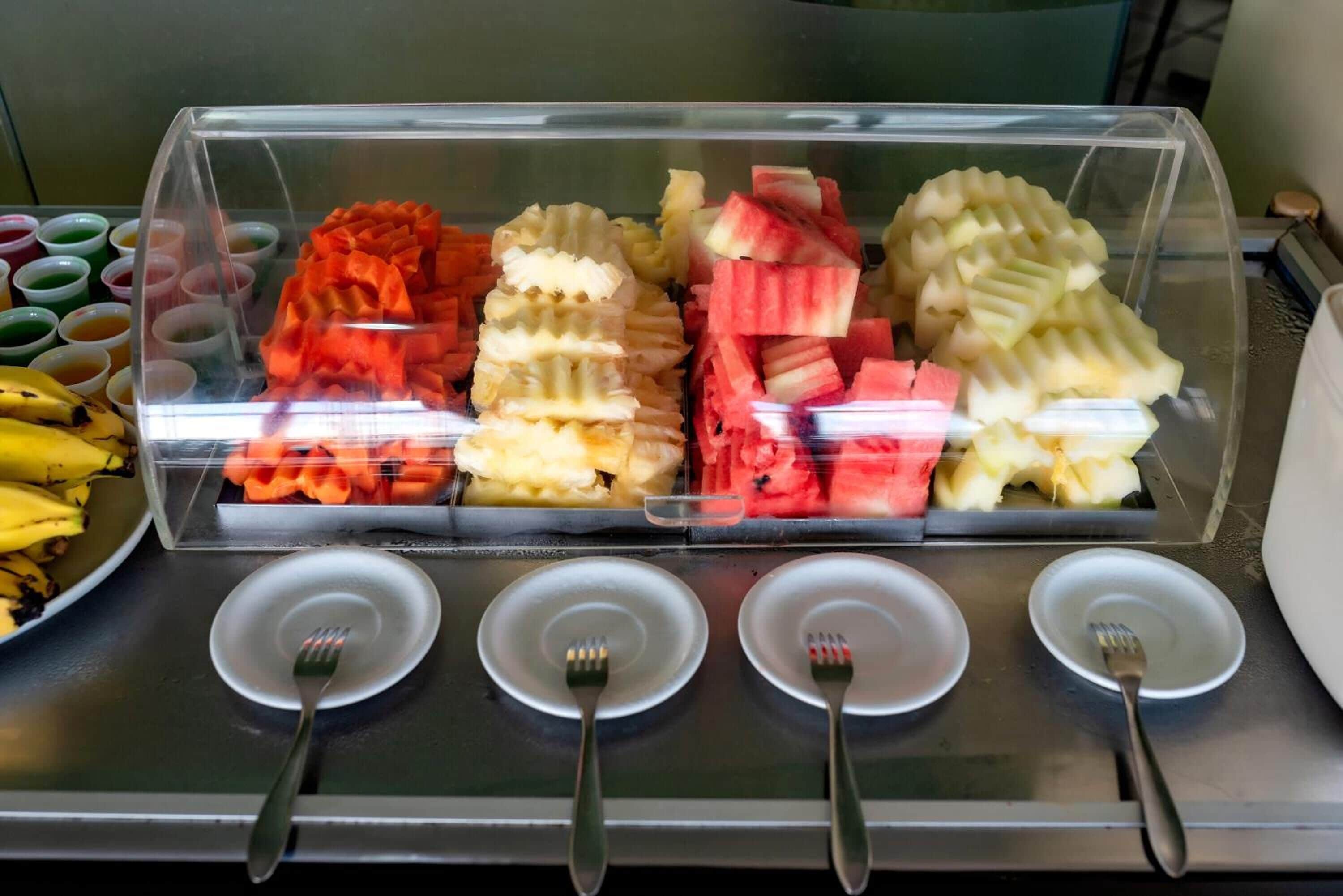 Une présentation de plats comprenant différentes sortes de fruits coupés en tranches et un plat de bananes, accompagnée de quatre assiettes vides et de quatre fourchettes.