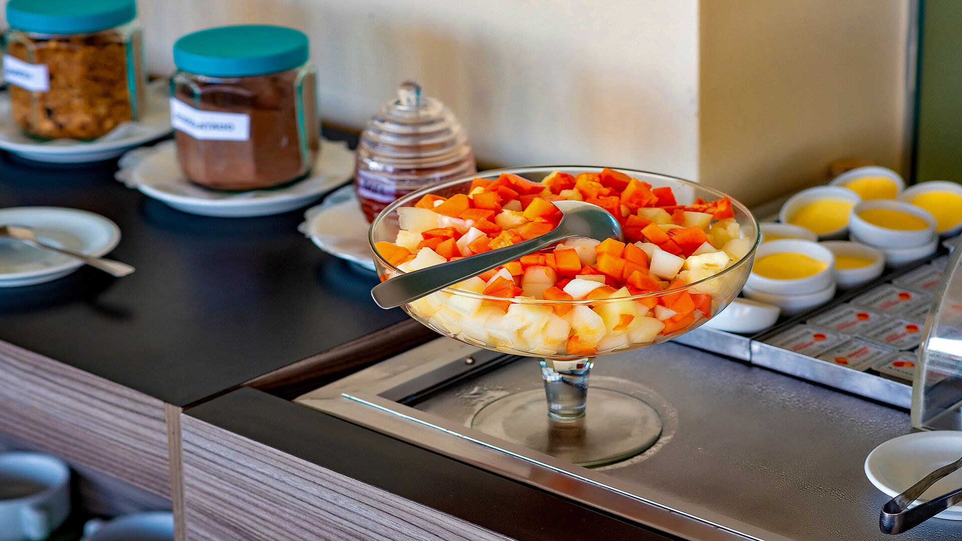 En buffet med en glas skål fyldt med frugtsalat, en honningske og forskellige tilbehør i baggrunden.