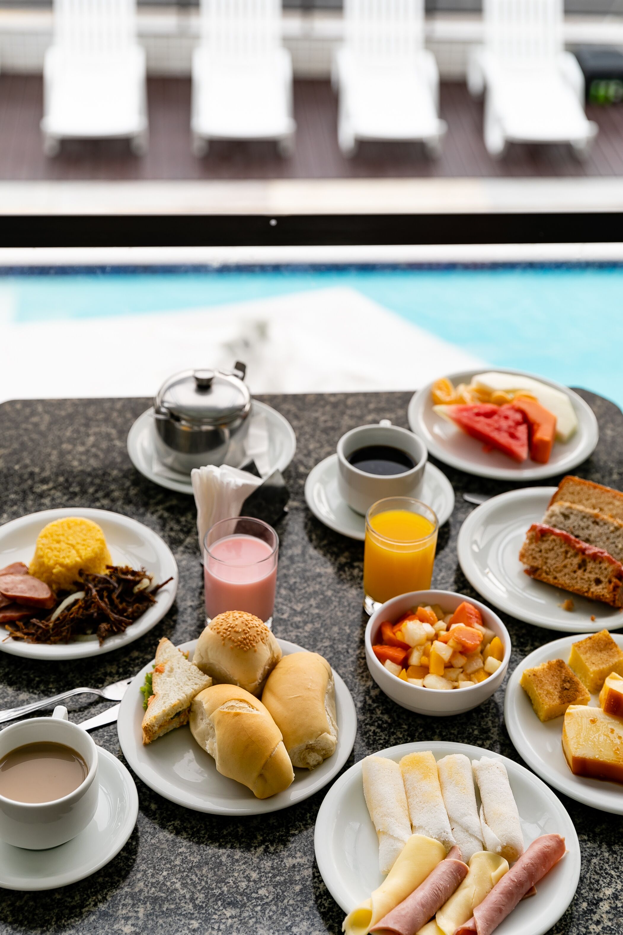 Una mesa con alimentos para el desayuno, como sándwiches, frutas y bebidas, con una piscina y sillas de descanso al fondo.