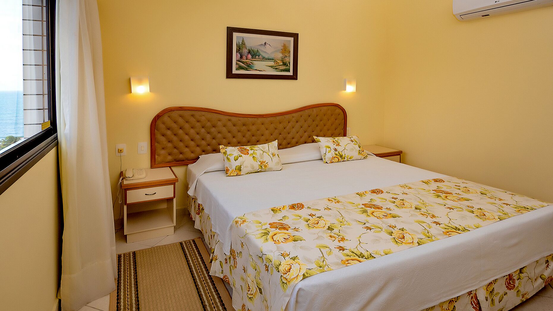 A hotel room with a bed, a nightstand, a lamp, a painting on the wall, and a window with blinds.